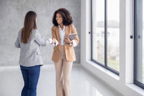 Two women shaking hands in a bright office space, symbolizing the beginning of a professional relationship. One woman holds a notebook, suggesting they are engaged in a business discussion. This image could represent the next steps in a real estate career after obtaining a license.